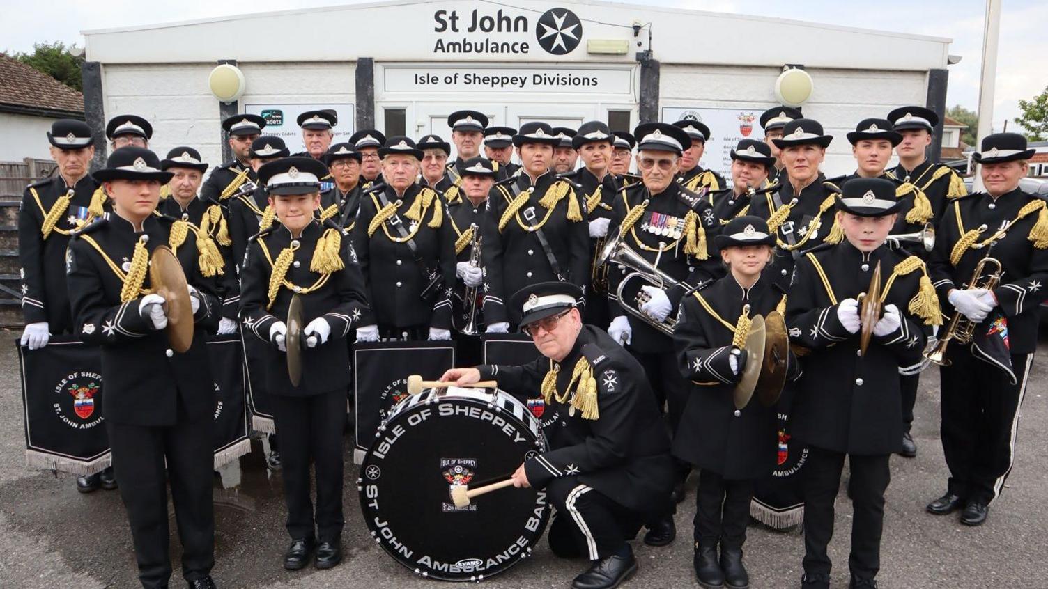 St John Ambulance band members dressed in black uniforms, with black caps and gold braiding on their shoulders standing in front of a white building.