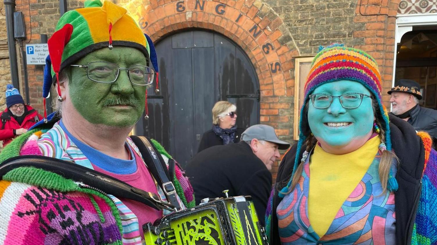 A man and woman in colourful clothing both wearing hats are looking at the camera and smiling. The man on the left has a green painted face and is stood next to a woman who has a blue painted face.