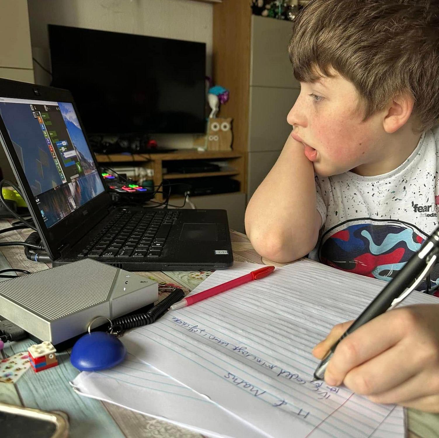 A young boy with brown hair wearing a white t shirt. he is sat at a desk with a pen in his hand and papers in front og him, looking at a laptop. His head is in his hand and he is looking at a black laptop with a fed-up expression on his face.