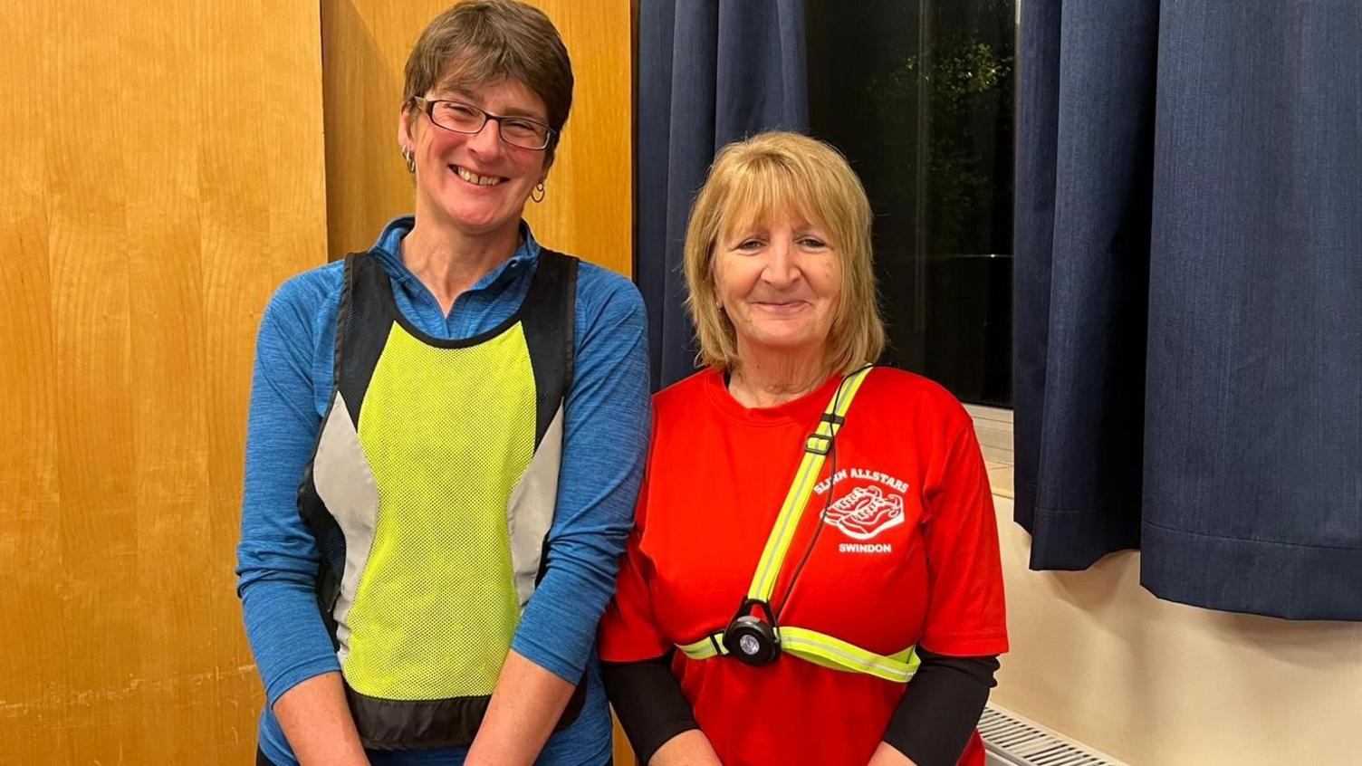 Two women standing indoors in a brightly lit room. The woman on the left is wearing a long-sleeved blue top with a neon yellow reflective vest and a black waist belt, along with a sports watch. The woman on the right is wearing a bright red T-shirt with a printed logo reading “Slinn Allstars Swindon” layered over a black long-sleeved top. Both have head torches strapped across their chests. Behind them is a wooden panel and a window with dark blue curtains.