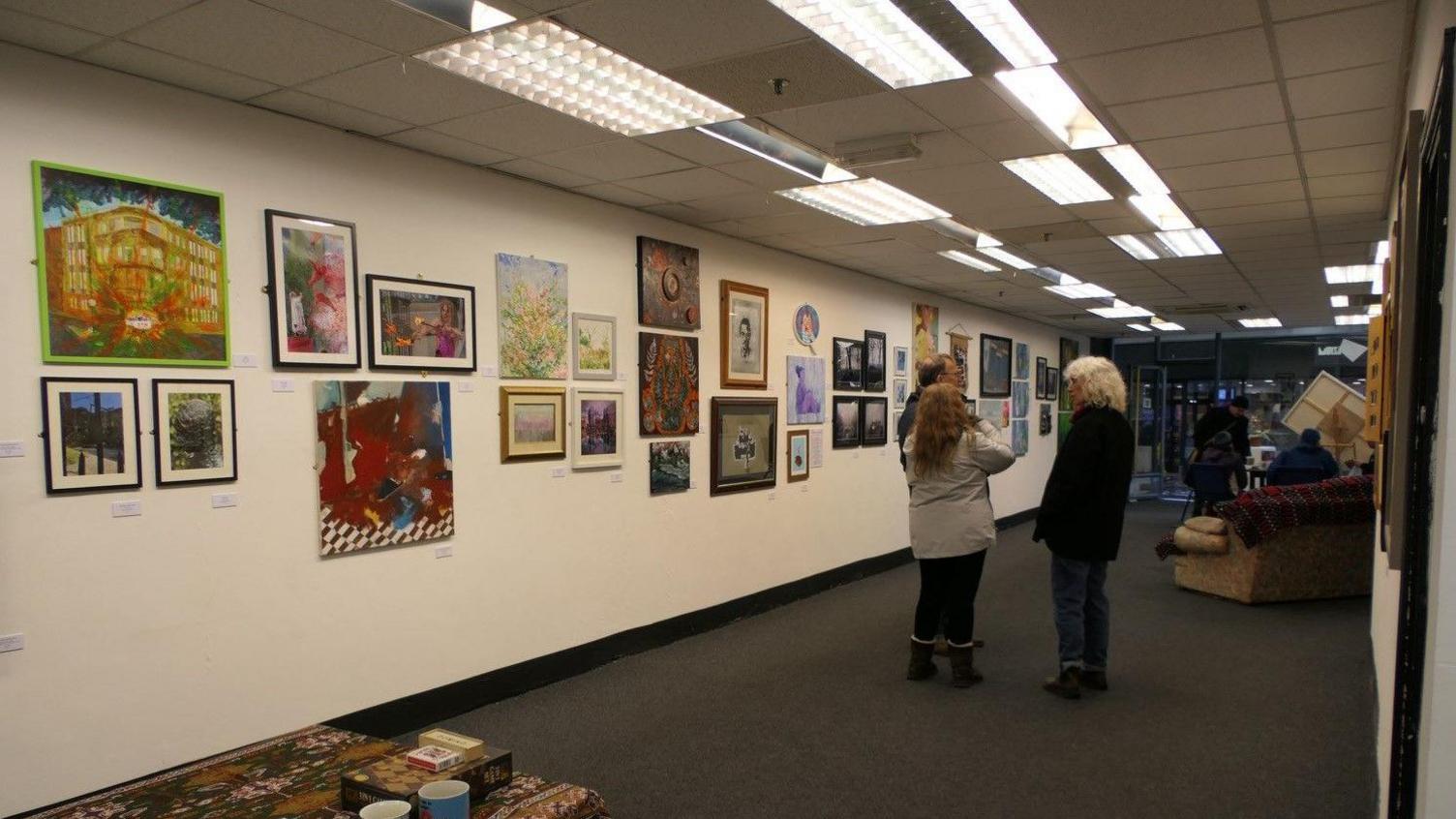 A long room with white walls and strip lighting in the ceiling with colourful paintings on one wall and a group of people standing talking to one another opposite.