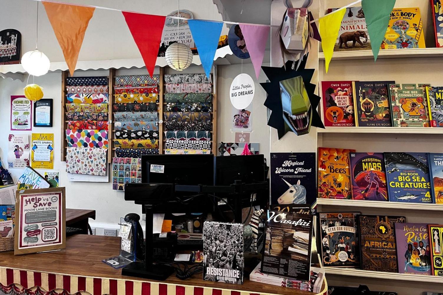 The cashier's desk at a book store. There is a dark brown desk, with a red and white striped decoration on the side. A computer screen is on right of the desk, surrounded by books including 'Resistance' and 'Magical Unicorn Friend'. There is a small gap on the desk to the left of the computer. To the left of this is a poster which reads 'Help us save Moon Lane Books', with a QR code. There is a display of wrapping paper on the white wall behind the desk, and multi-coloured triangle banners are displayed from the ceiling. To the right of the desk, there is a set of shelves with colourful children's books on display.