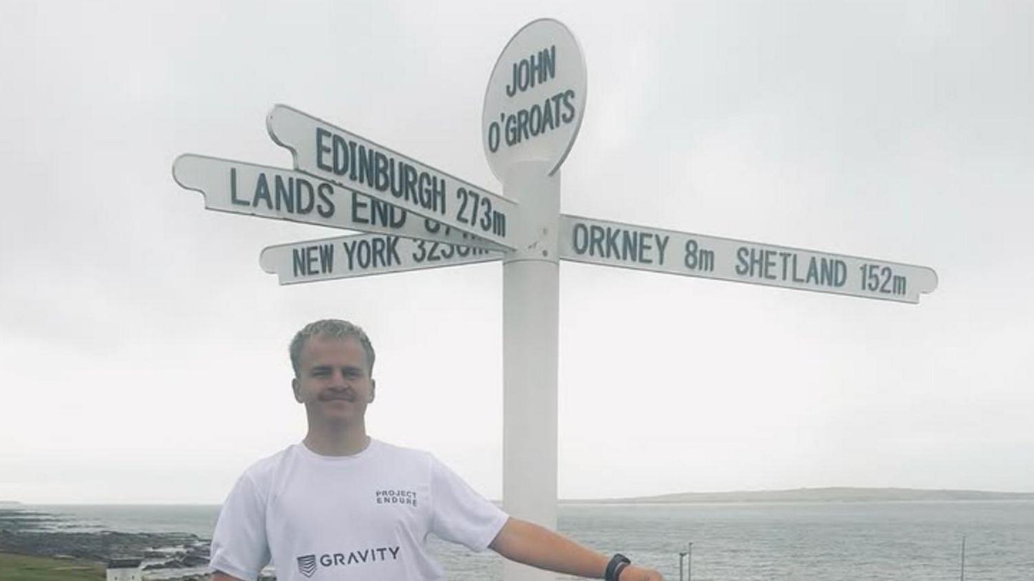 Drew Jonkmans is pictured dressed in a white T-shirt. He is a young man with short blonde hair and he is smiling. He is standing in front of a signpost marked John O' Groats. The sign has fingers pointing to Edinburgh, Land's End, New York, Orkney and Shetland. The sea can be seen in the background.