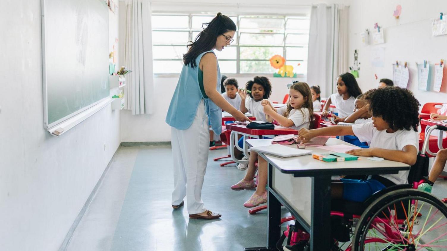 A classroom assistant wearing a blue top and white trousers helps children in a school with their learning.