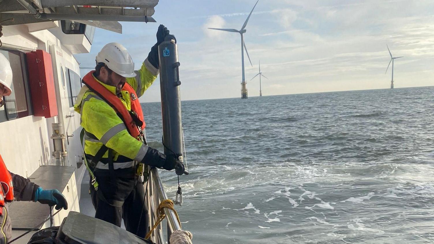 A man wearing a white hard hat and a yellow waterproof, looking up and looking at core sample, standing on a white boat, with the sea around him and wind turbines in the distance  