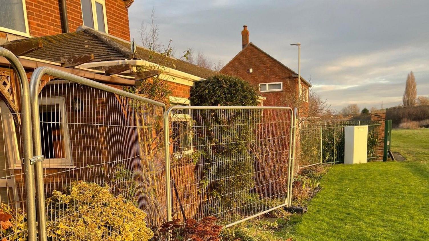 Heras fencing installed on the very edge of residents' homes. Their back gardens are on the left of the image, inches away from the fencing which blocks the view from their windows. On the right side of the fencing is a lush green field. 