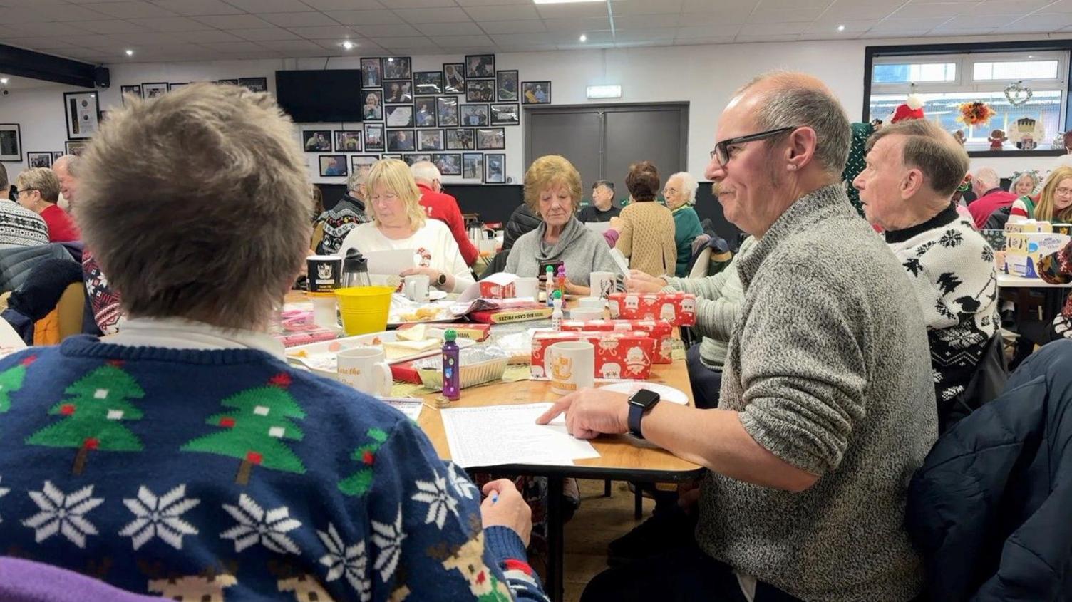 Men and women sit around a table, including wrapped presents and mugs, in the foreground. Other men and women are sitting down in the background.