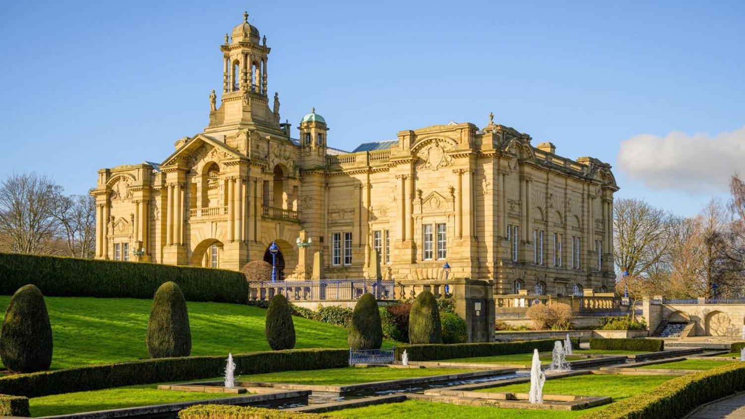 A grand building bathed in bright sunlight, with carefully manicured gardens and fountains in the foreground. The sky is blue with little cloud. 