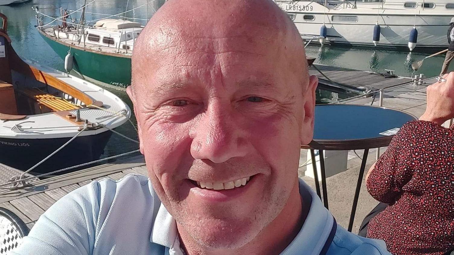 A bald man smiles at the camera. He is sitting next to a marina with small boats behind him