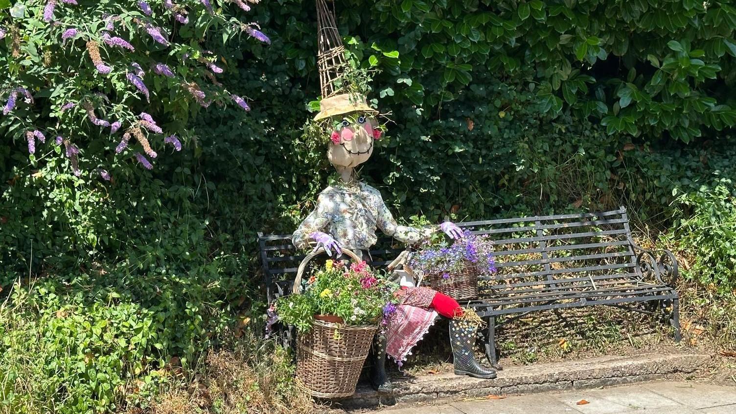 A scarecrow made to look like a woman in a shirt and skirt is sitting on a bench holding baskets with flowers. There are bushes around the bench.