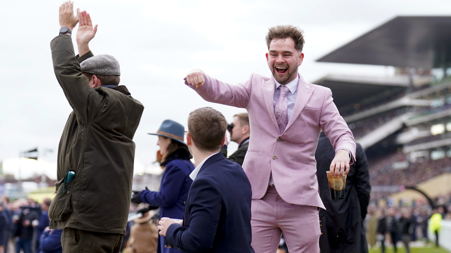 Racegoers are cheering during a race. On the left is a man in a dark green Barbour jacket clapping with his hands above his head. In the centre is a man in a dark blue blazer looking up at a man who is facing the camera, cheering, in a full lilac suit. He is holding the top of a pint cup.