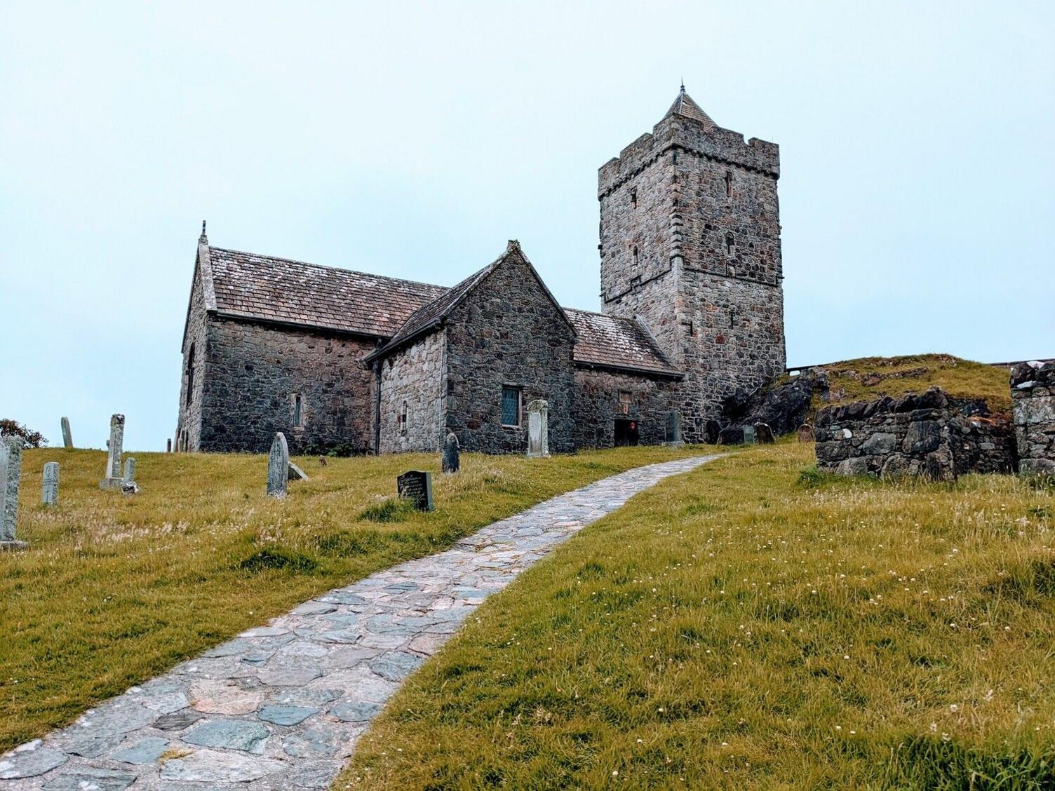 An old grey stone church with a couple of old grey headstones on a hill with yellowed green grass. A rocky path leads down from the hill to the viewer.