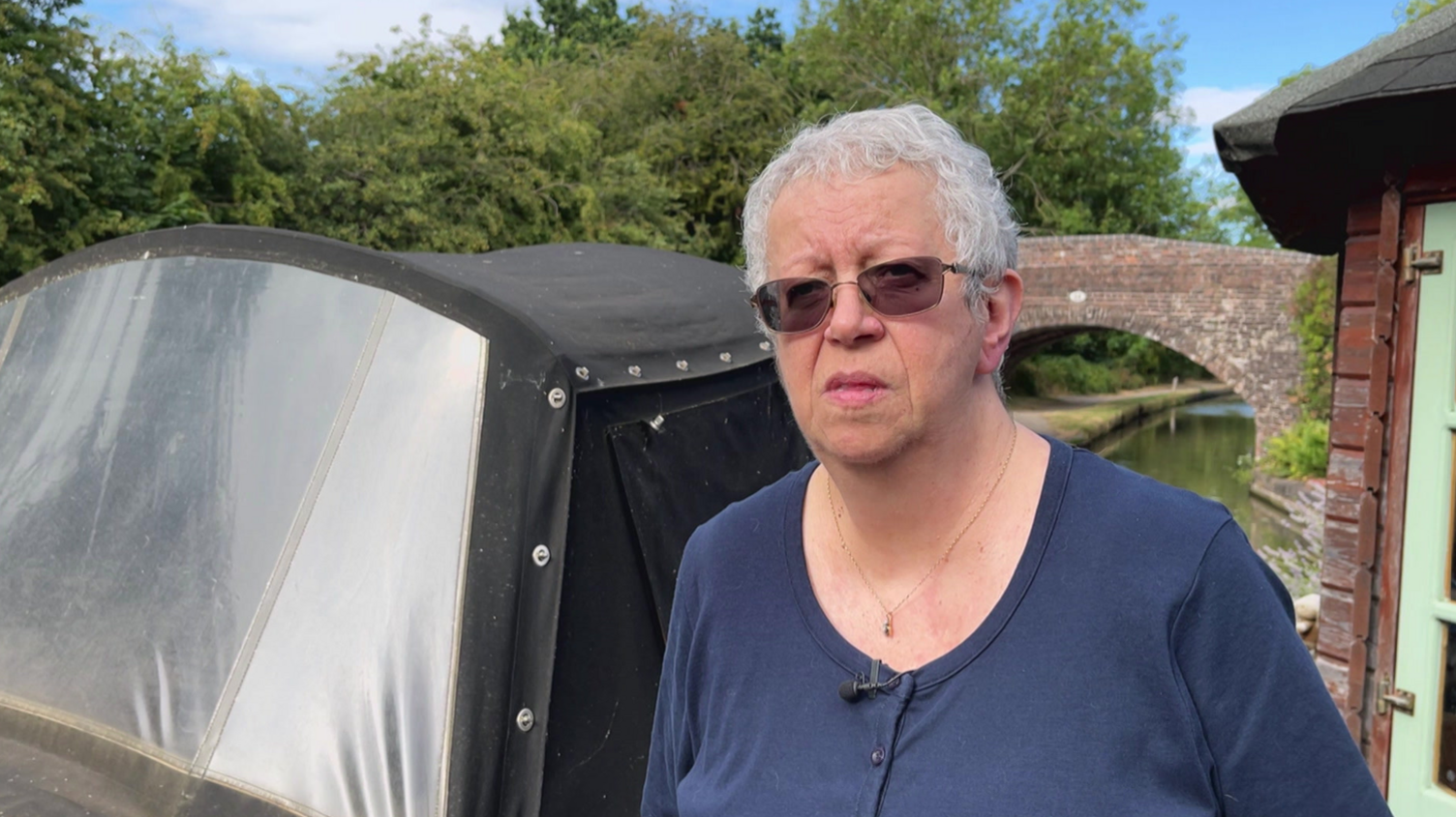 A woman with short grey hair, a dark blue top and sunglasses standing in front of a narrowboat