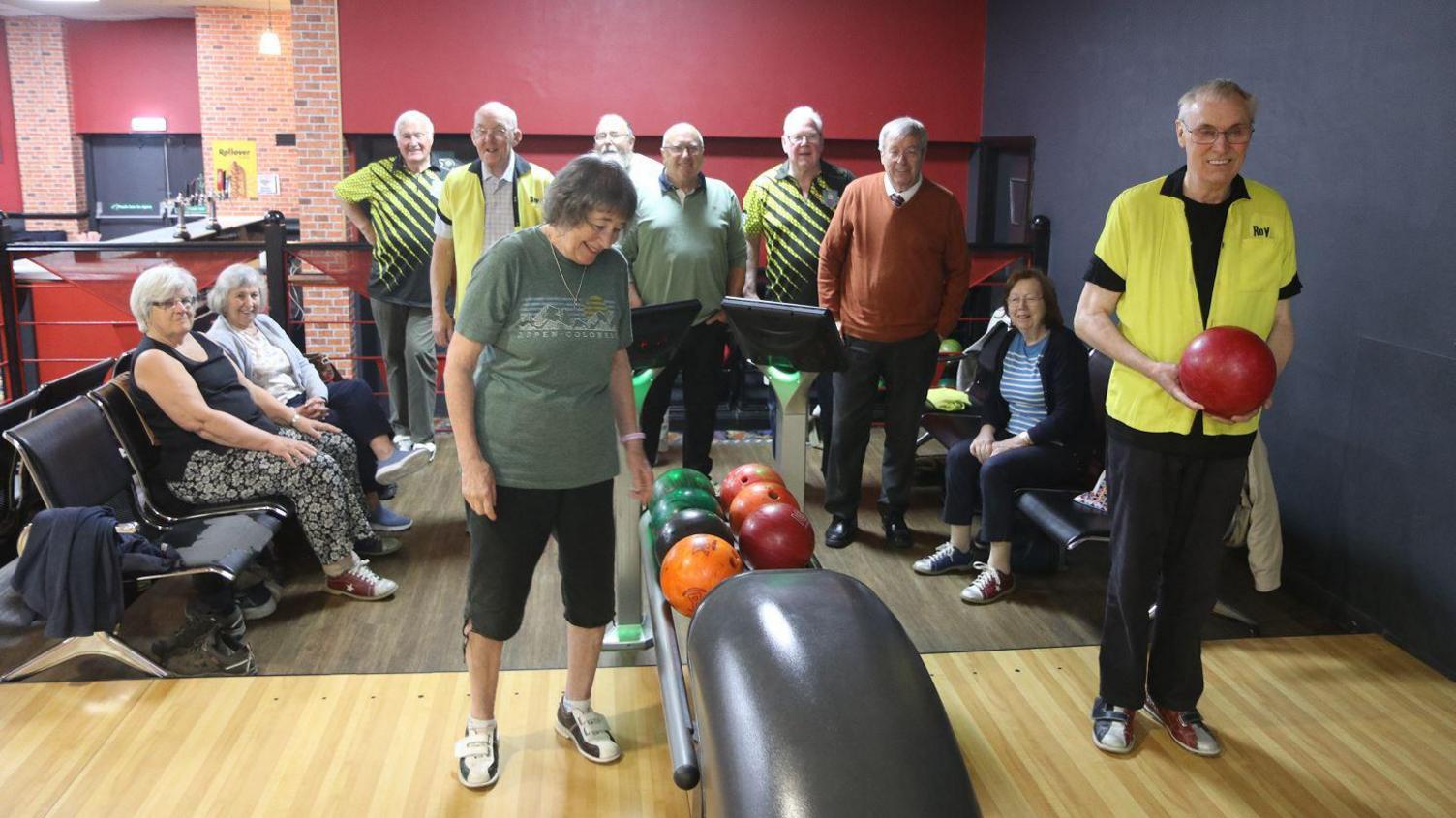A group of retired people playing ten pin bowls, one man is wearing a yellow t-shirt and holding a bowling ball.