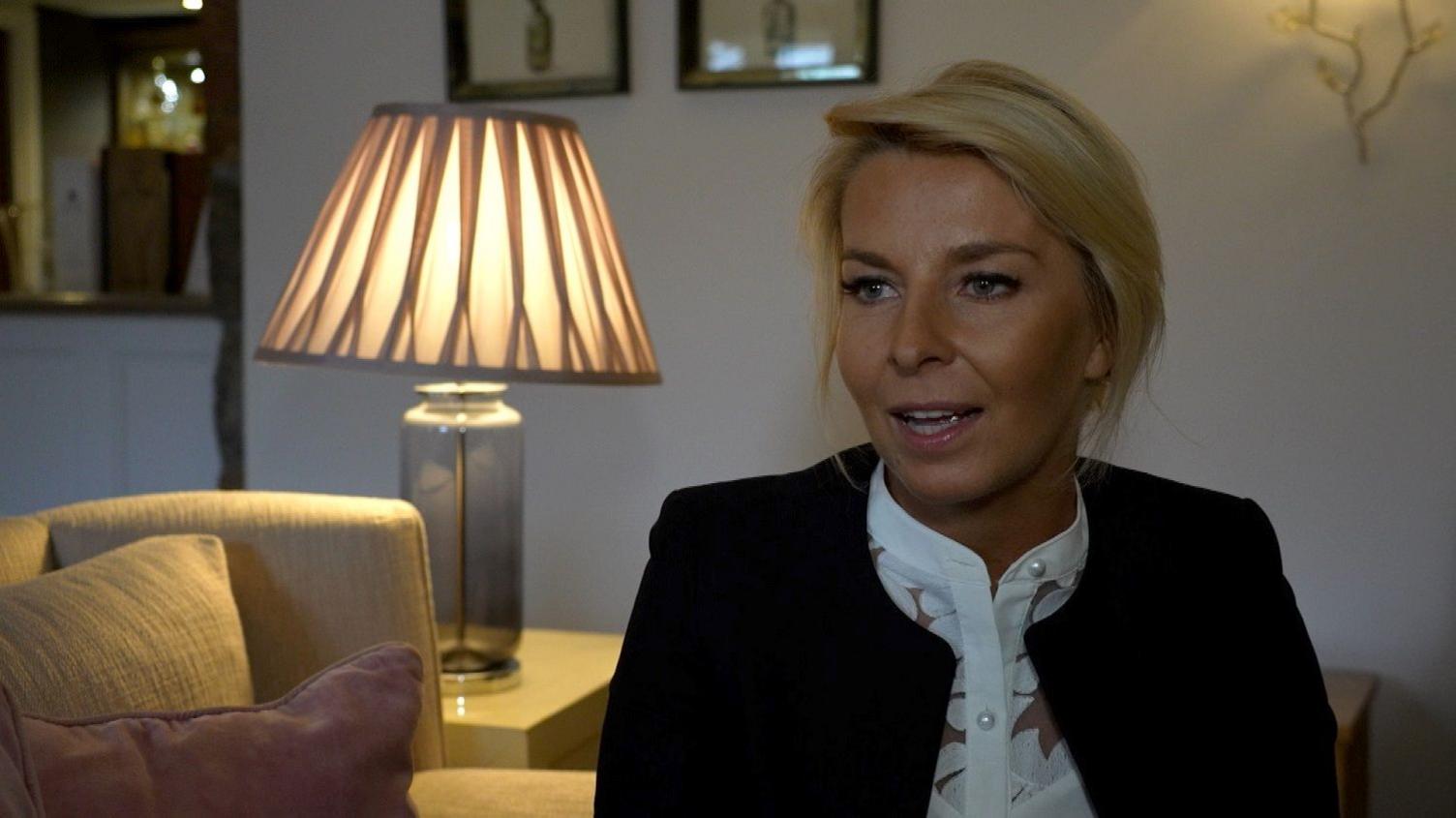 Ola Przyjenska seated a well-lit room or office space. She has blonde hair styled back and is wearing a white blouse with a dark jacket over it. 