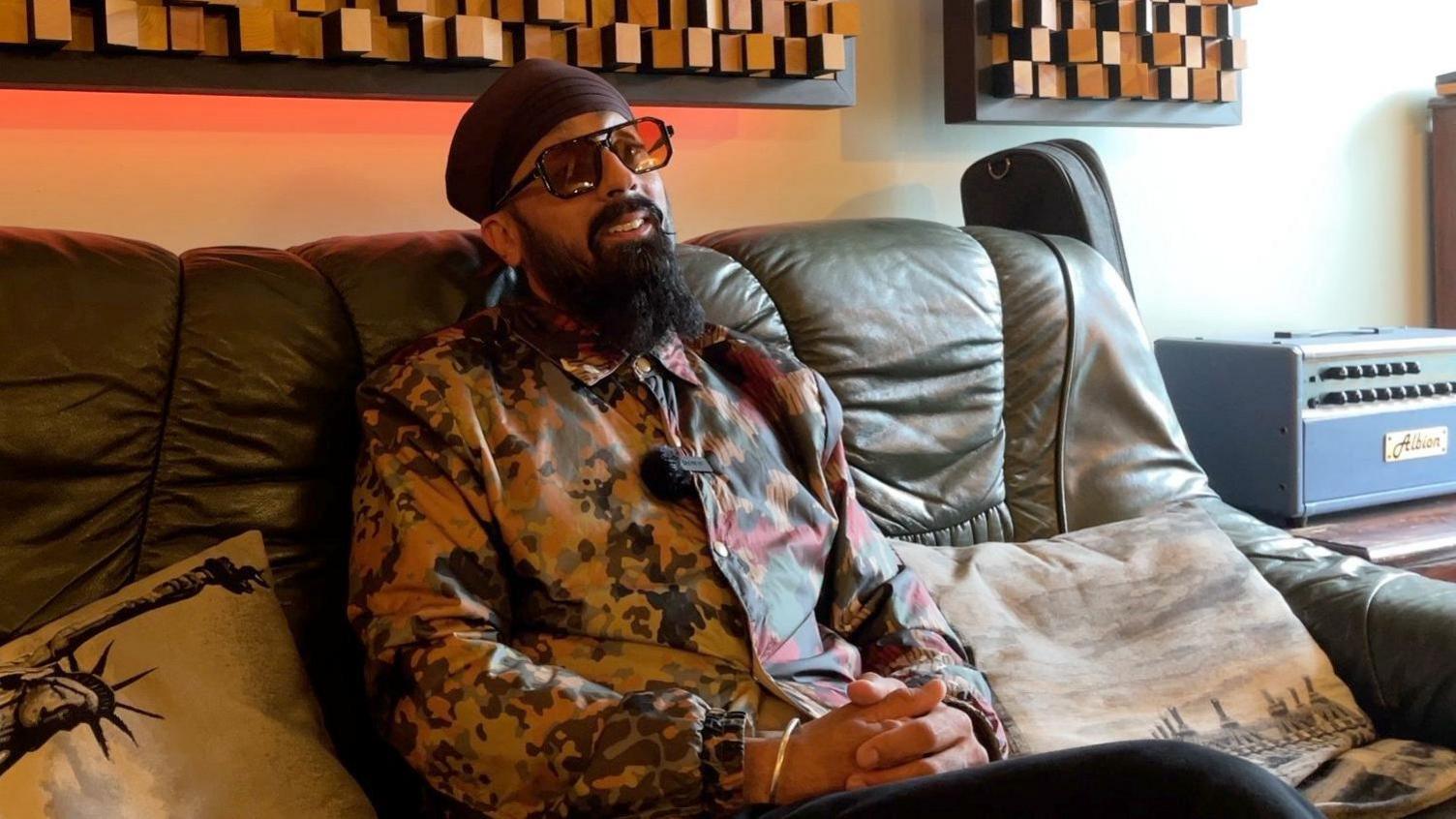 Silverfinger Singh sitting on a music studio sofa, wearing a cameo print jacket and big sunglasses. 