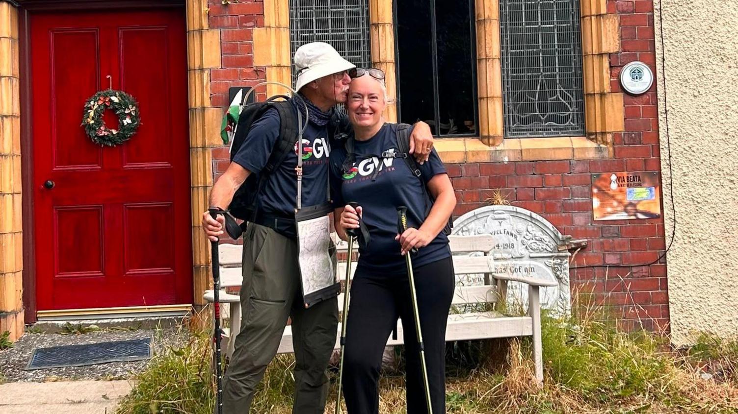 John kisses Hayley on the forehead, with his hand around her shoulder. They hold walking sticks, and stand in front of a red brick house with a red door.