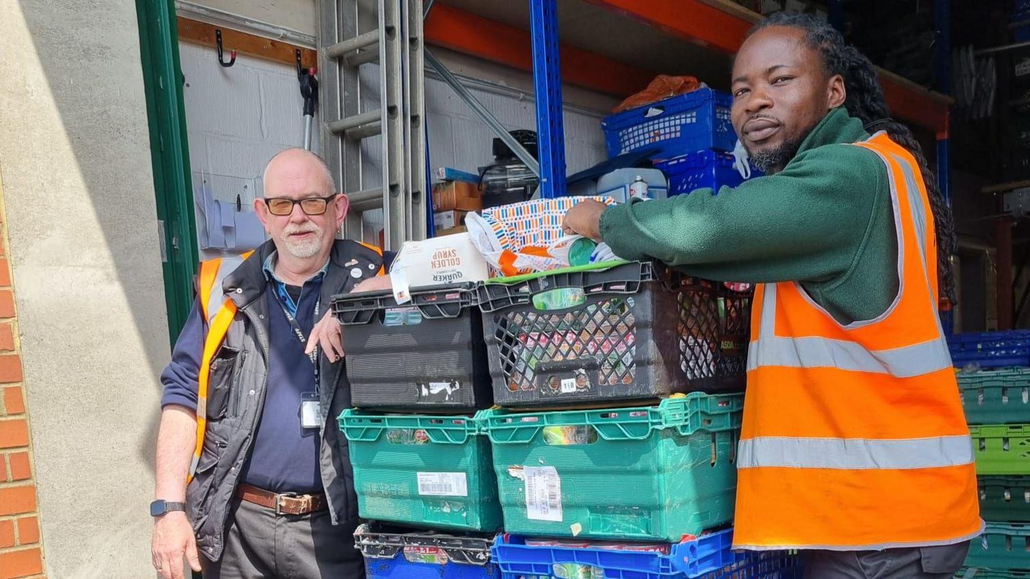 Two people standing by a large amount of boxes with food items in them. The boxes are black, green and blue. One man to the left is hearing a blue top, jacket, high-vis and trousers, the other man to the right is wearing a green top, dark trousers and a high-vis jacket. They are both looking at the camera.