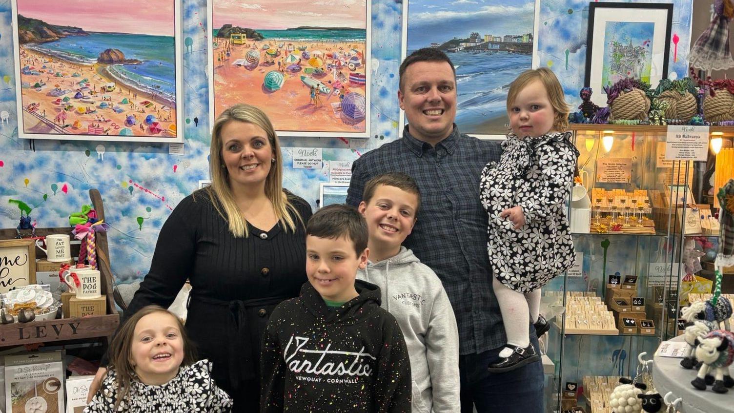 Rachel in a black top with her husband in a navy green checked shirt. He is holding a toddler girl and their three other children stand in front of the couple - two boys and one girl. Some of Rachel's paintings of the beach are displayed on the wall behind them