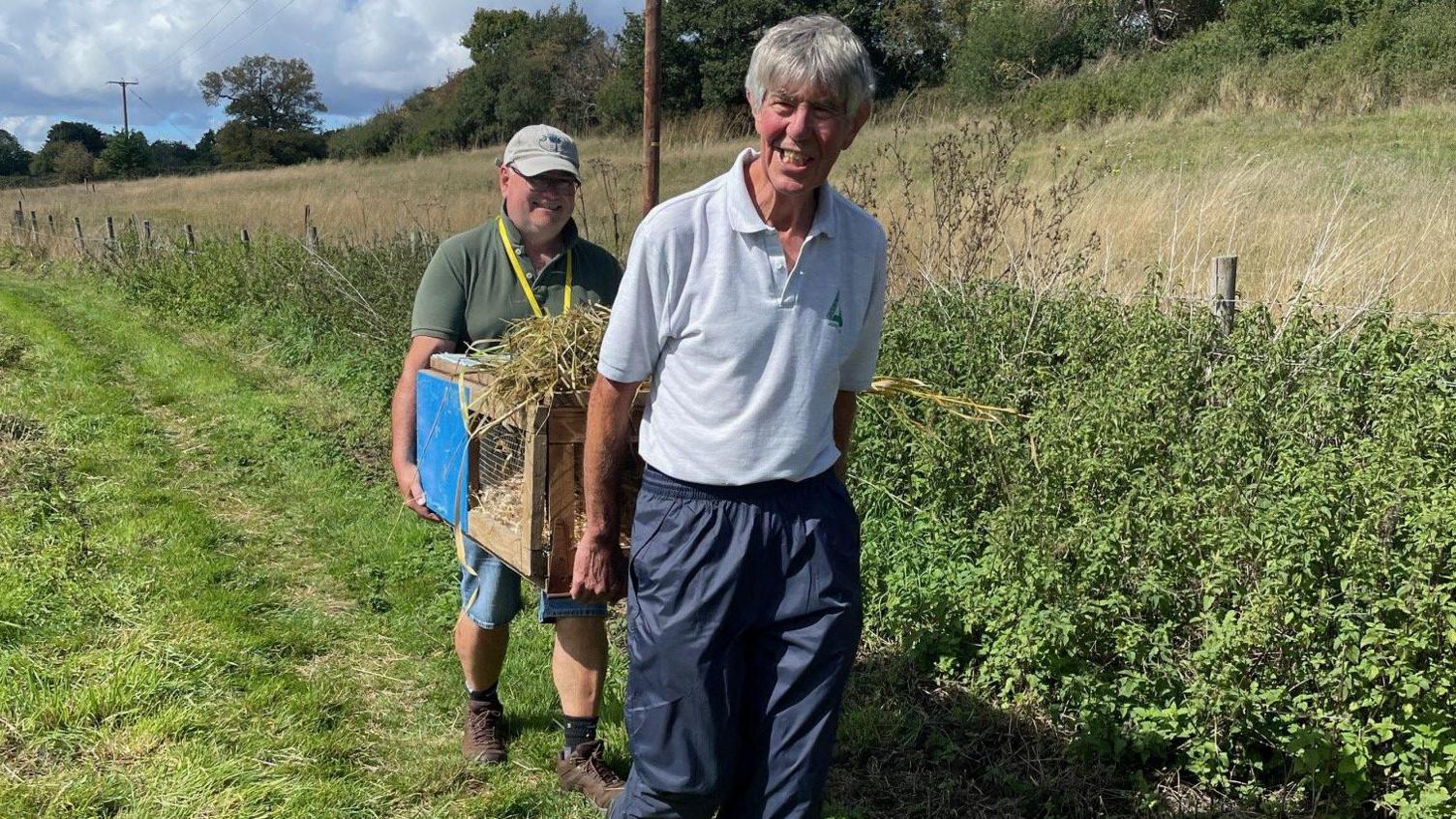 Two people, in a field, walking a water vole box, with a water vole in it, to a riverbank. There are trees and foliage around them. One is wearing shorts and a cap and the other man, at the front, has on a top and blue trousers. 