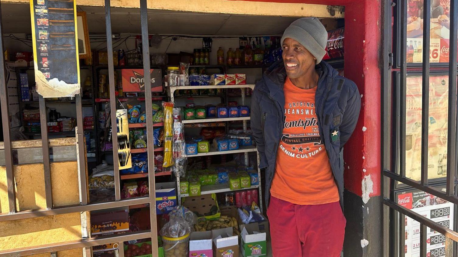 Julius Koobetseng in red trousers, oranage T-shirt, blue coat and a beanie hat stands outside his small shop. Shelves of goods can be seen in the background. 
