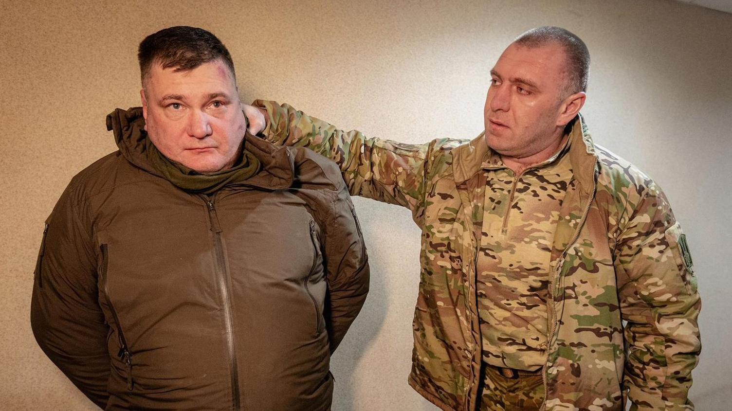 Ukrainian SBU chief Vasyl Malyuk (right) stands with his hand behind an arrested man's head, apparently holding him by the hood of his jacket. Malyuk is  dressed in military-style fatigues and the other man is wearing a zipped-up brown jacket.