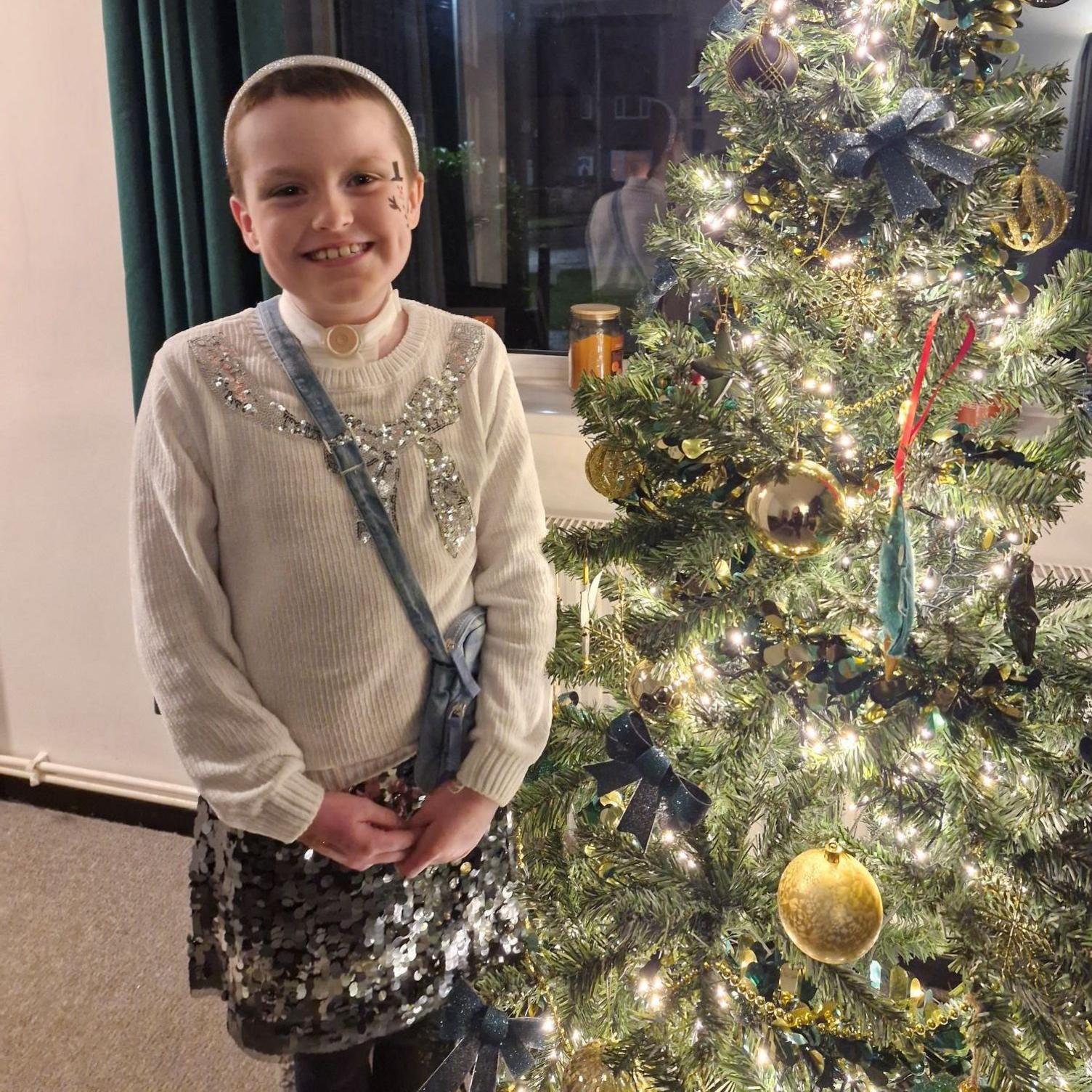 A girl is standing next to a Christmas tree. She is wearing a silver sequinned skirt and a cream jumper with sequins embroidered on it in a bow pattern. She has very short brown hair and is wearing a silver headband.