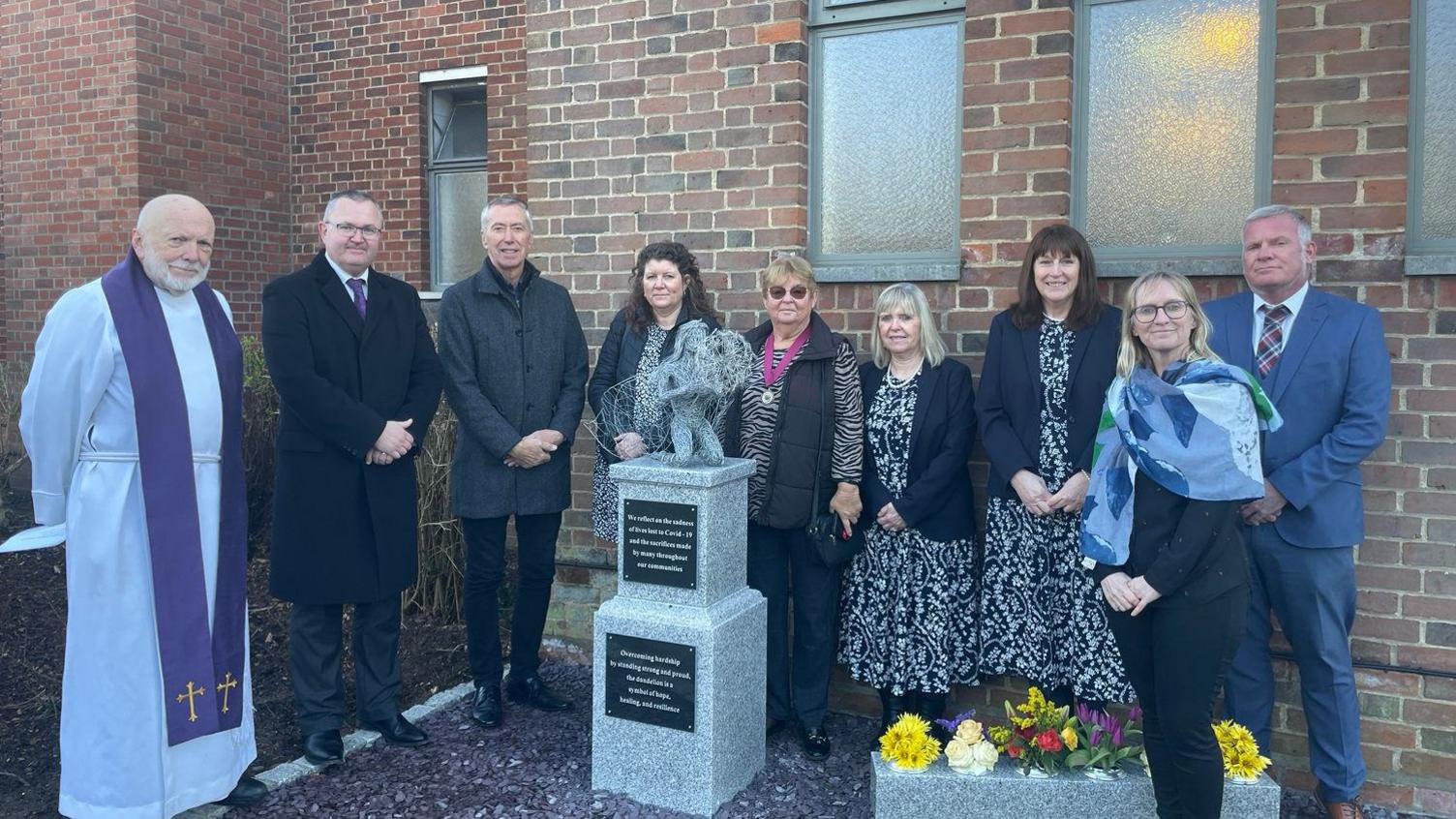 Nine people, including a vicar, stand around the Covid-19 memorial. They are looking at the camera and smiling. The memorial consists of two rectangular marble plinths, one on top of the other. It is topped with the figure of a kneeling woman holding a dandelion. Behind the gathering, the side of a brick building can be seen.