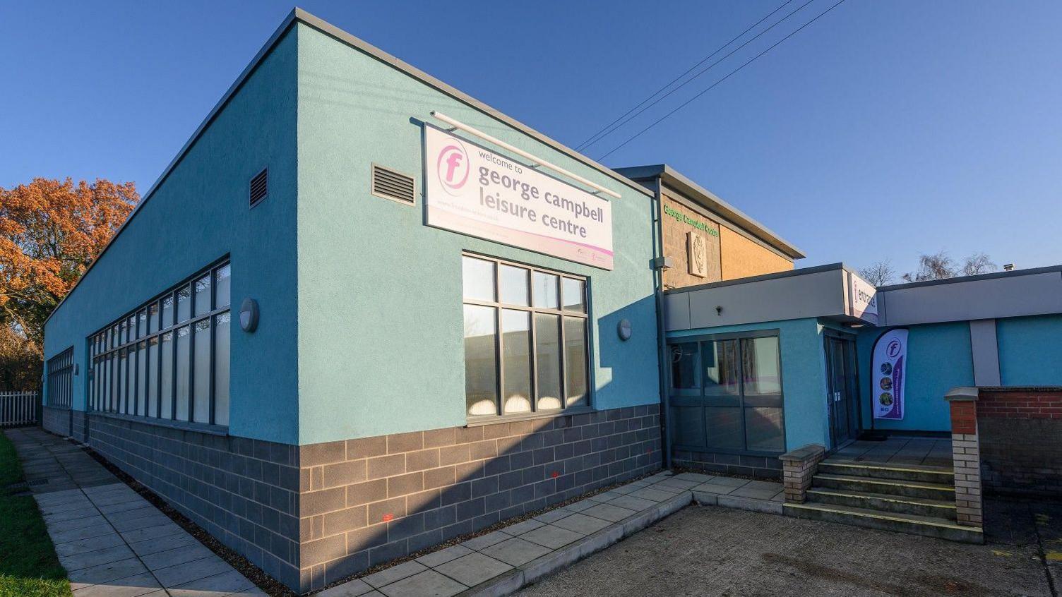 The George Campbell Leisure Centre is a modern looking building with light blue exterior walls, with breeze block visible from about 1m (3ft) up from the ground. There is a flight of four steps to the right hand side leading to some glass double doors. It has a large sign on the top facade with its name on.