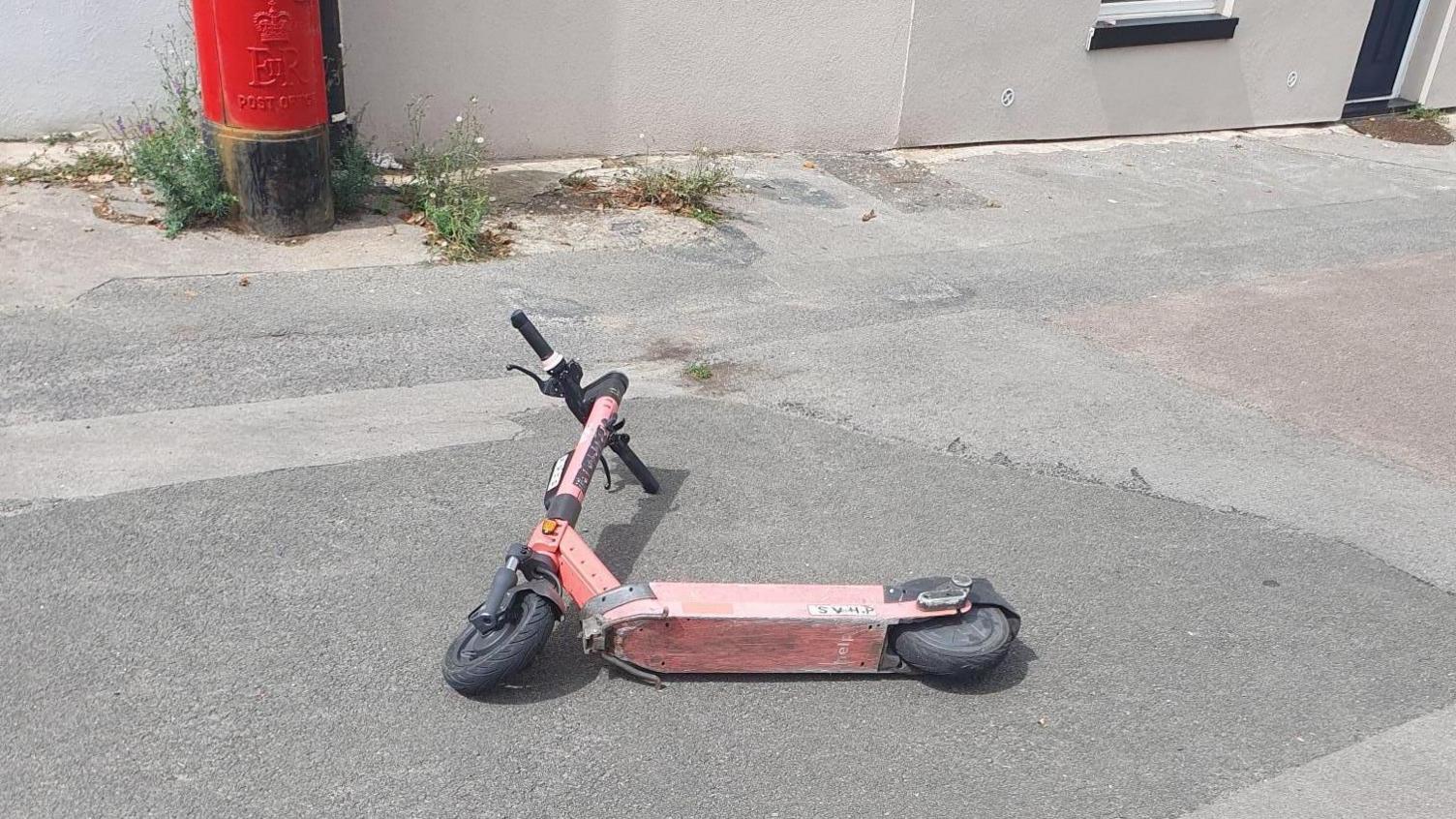 A faded red Voi e-scooter laying on its side in the middle of a pavement. It has been abandoned by a user.