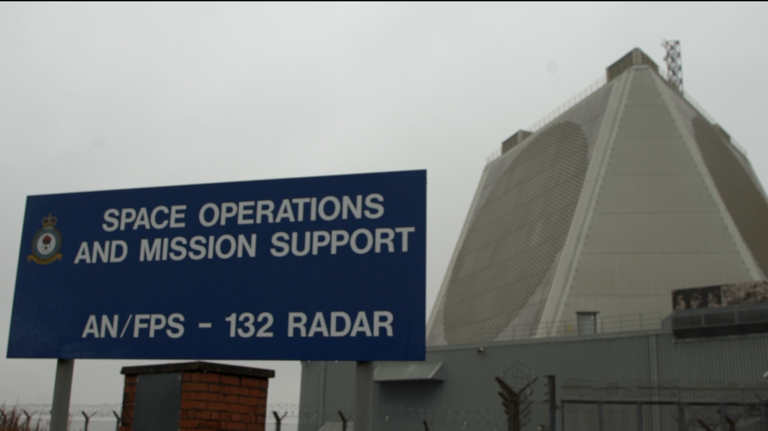 The exterior of the air force base. It's a tall, grey structure surrounded by small huts and barbed-wire.