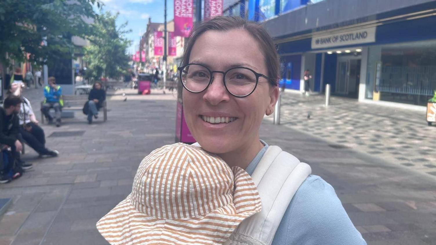 A woman carrying a baby. She is wearing a blue jumper and glasses and the baby has on a striped hat.