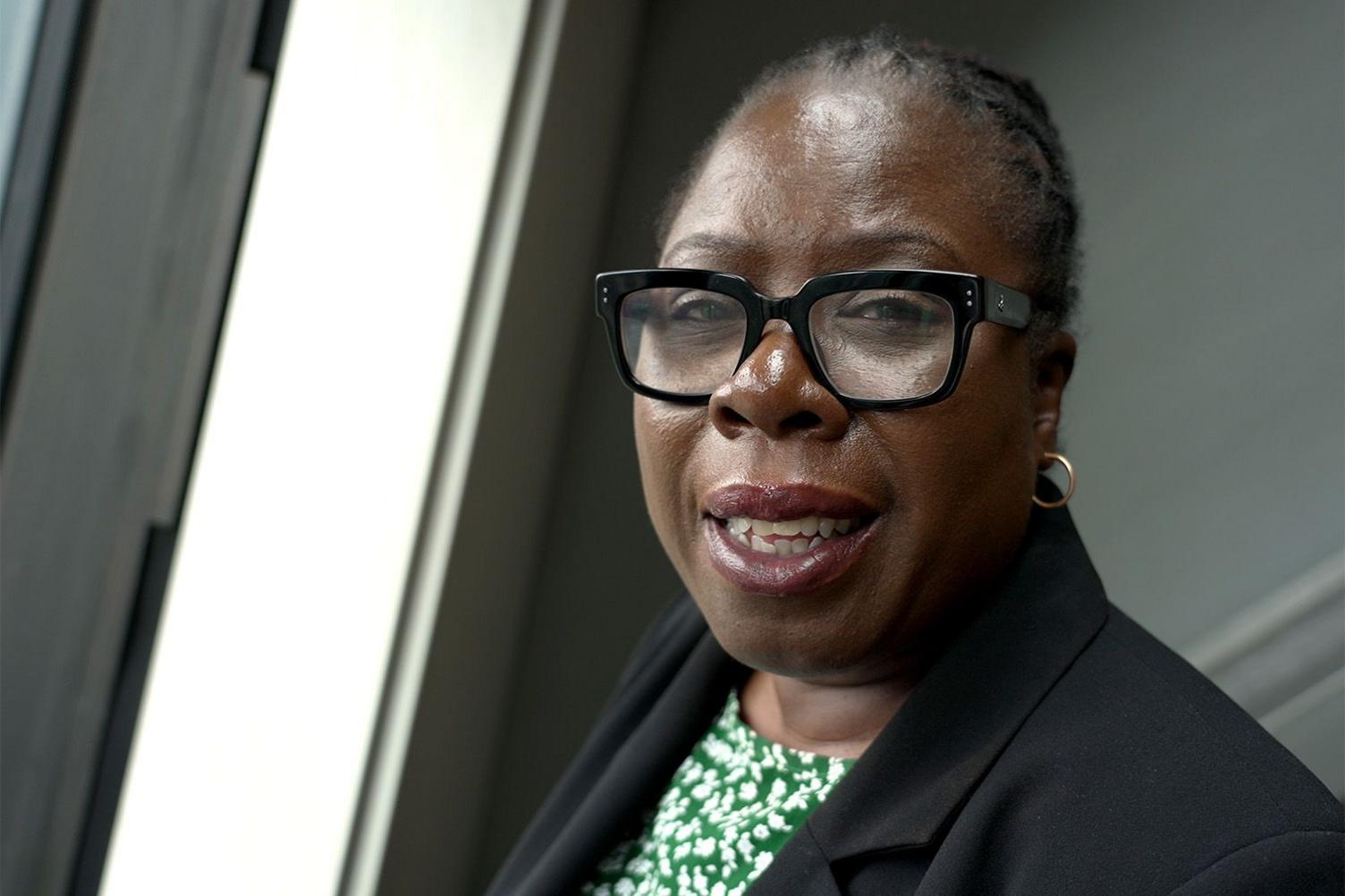 A headshot of Labour MP Paulette Hamilton. She is wearing thick rimmed black glasses, has small gold hoops in her ears and is wearing a black blazer over a patterned green and white top.