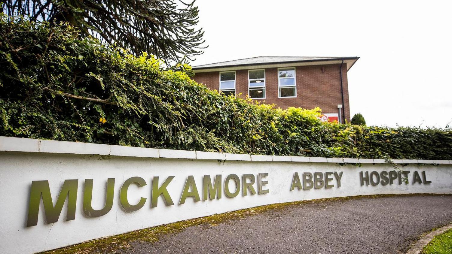 A sign says Muckamore Abbey Hospital on a low white wall in front of bushes and a red-brick building