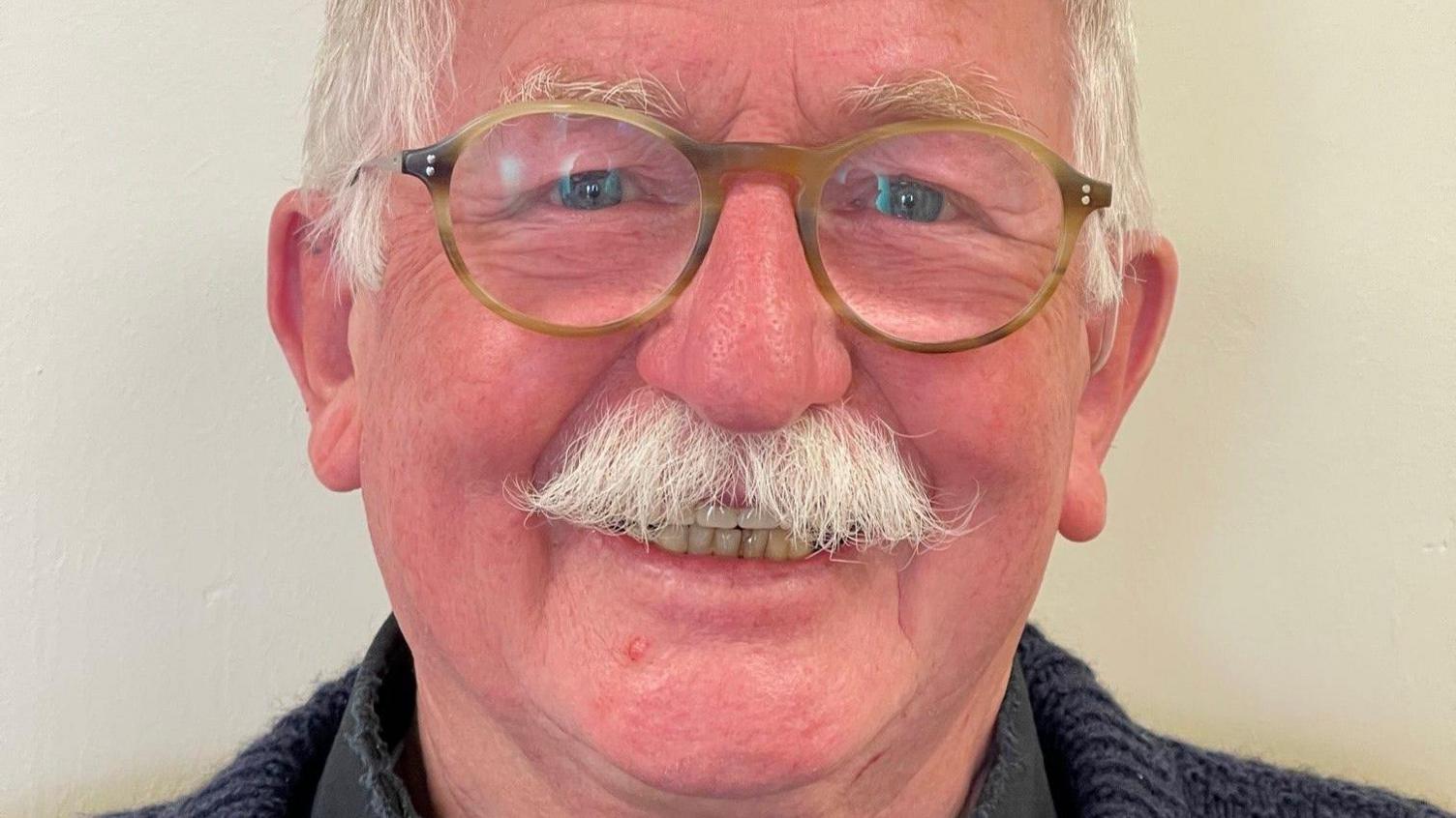 Councillor Bernie Fisher. He is smiling. He has white hair and a moustache. He wears rounded glasses, and a navy knitted jumper.