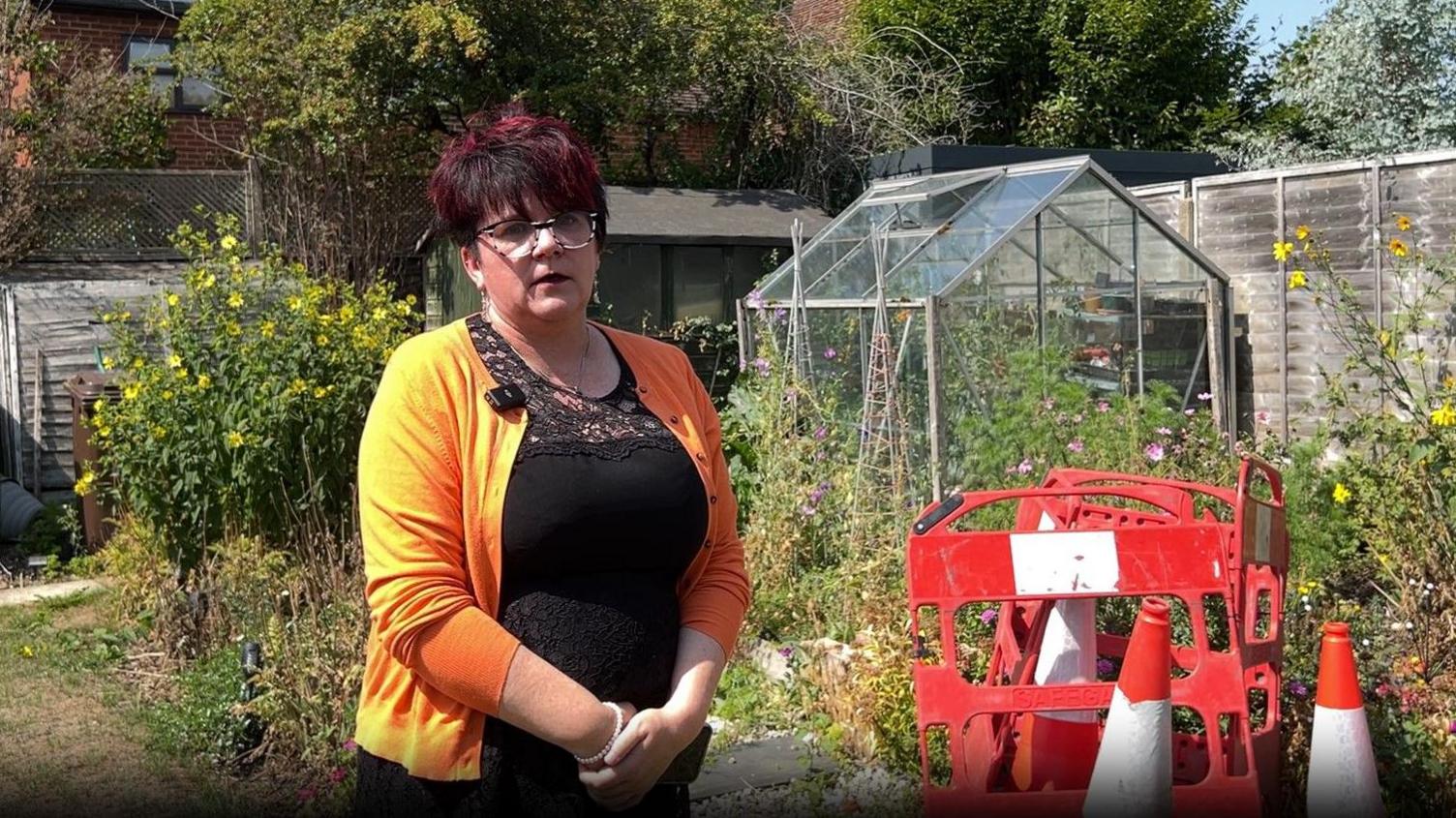 A woman stood next to some red cones and barriers in her garden next to some plants and a green house