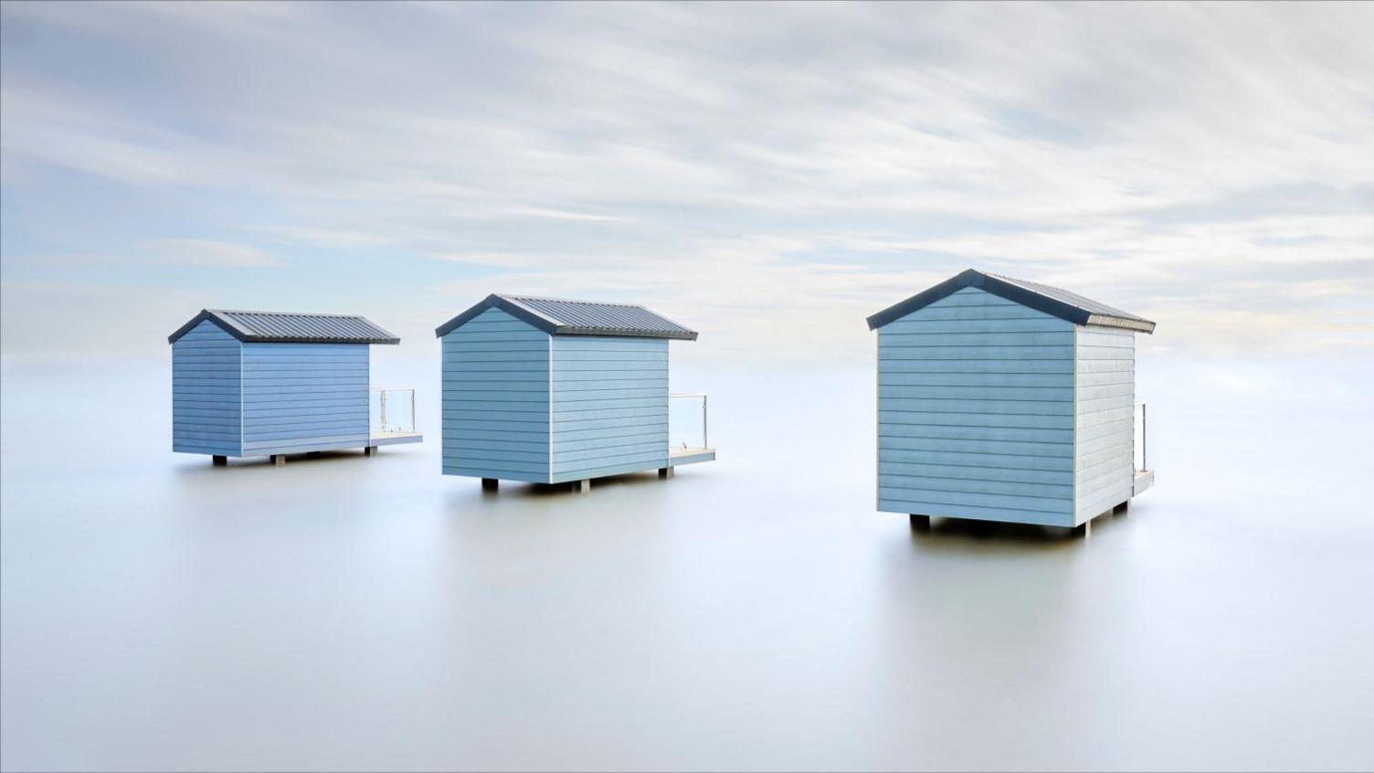 Three blue beach huts are standing in what appears to be an empty plain.