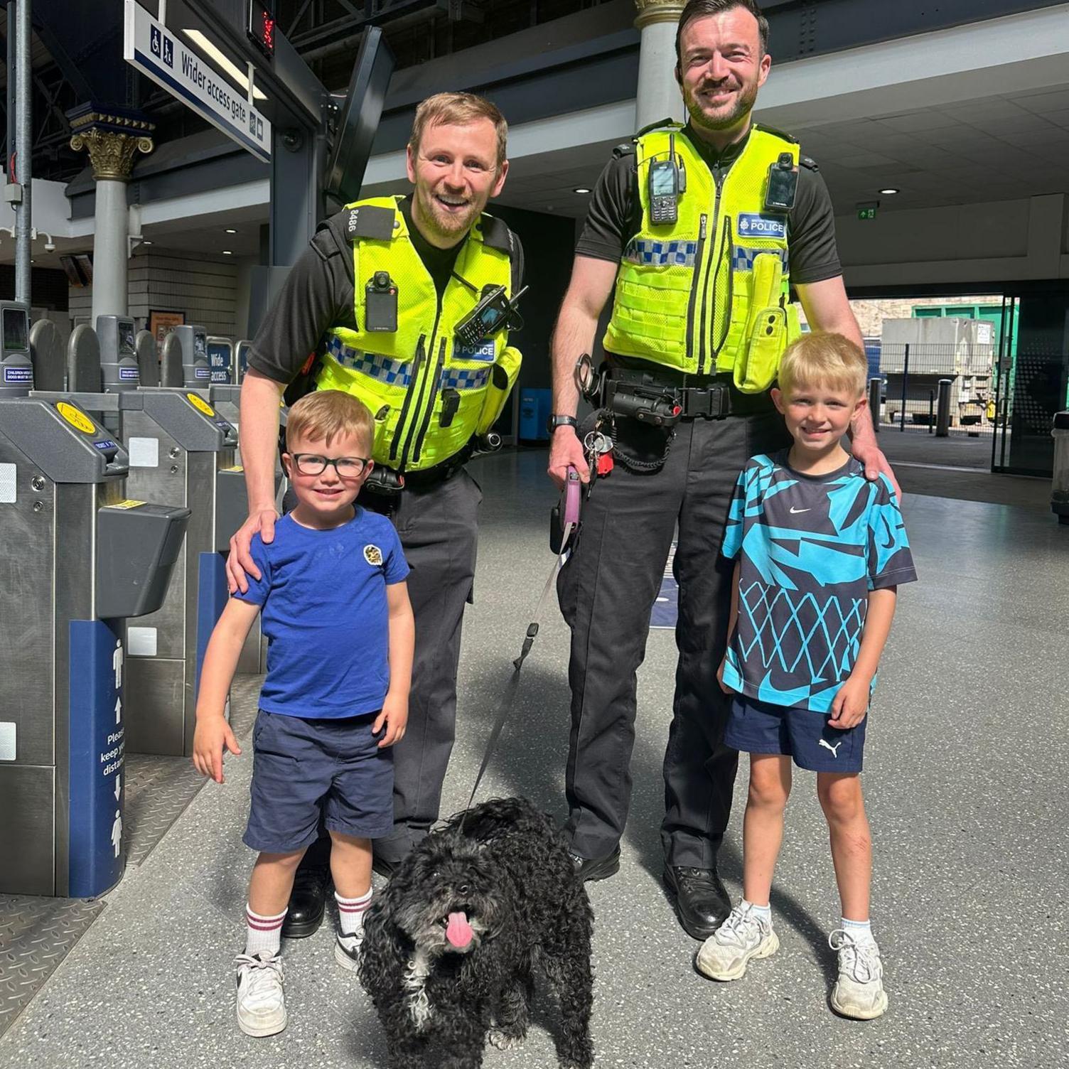 Two male police officers standing with two young boys holding a black dog on a lead.