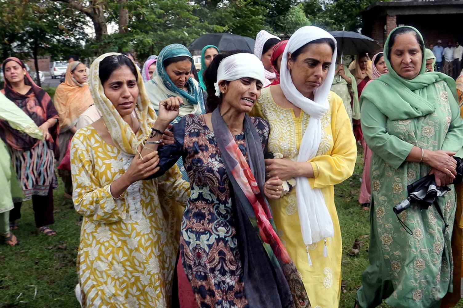 A group of women dressed in traditional clothing and headscarves, standing outdoors with trees and a building in the background. The woman in the centre - Vihaan's mother - is wailing.
