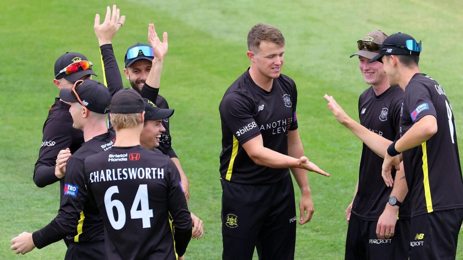 Gloucestershire cricket club players celebrate taking a wicket against Leicestershire. The players are in a huddle and wearing their dark one-day kit with yellow emblems