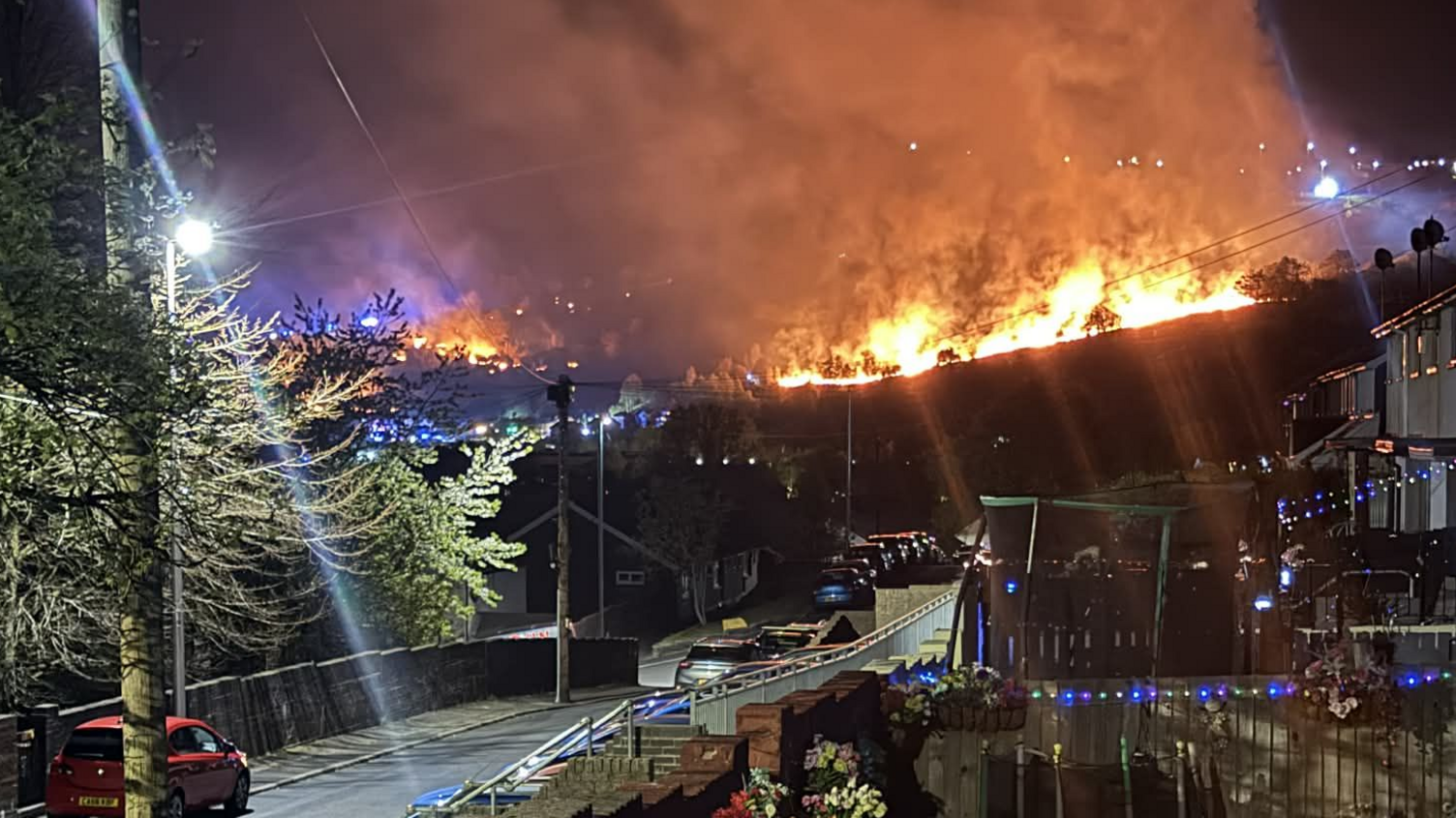 Llun gyda'r nos o fflamau ar fryn yn y cefndir ger rhes o dai 