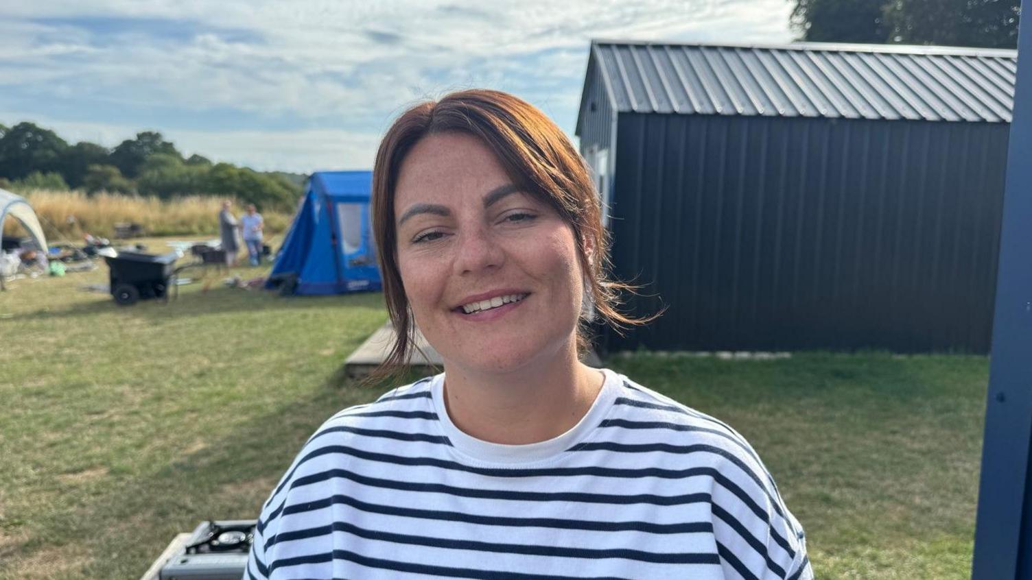 A woman with a white T-shirt with black horizontal stripes is smiling looking at the camera. There is a building to her right on the photo and a light blue tent in the middle is blurred.