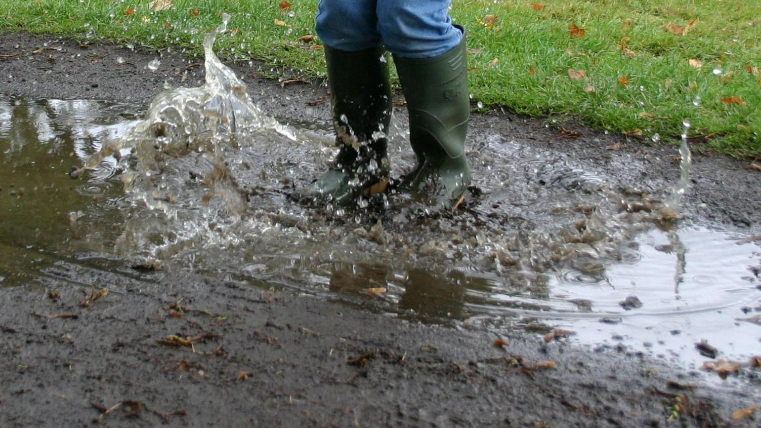 A picture of a child wearing wellies jumping into a muddy puddy. The wellies are dark green in colour.