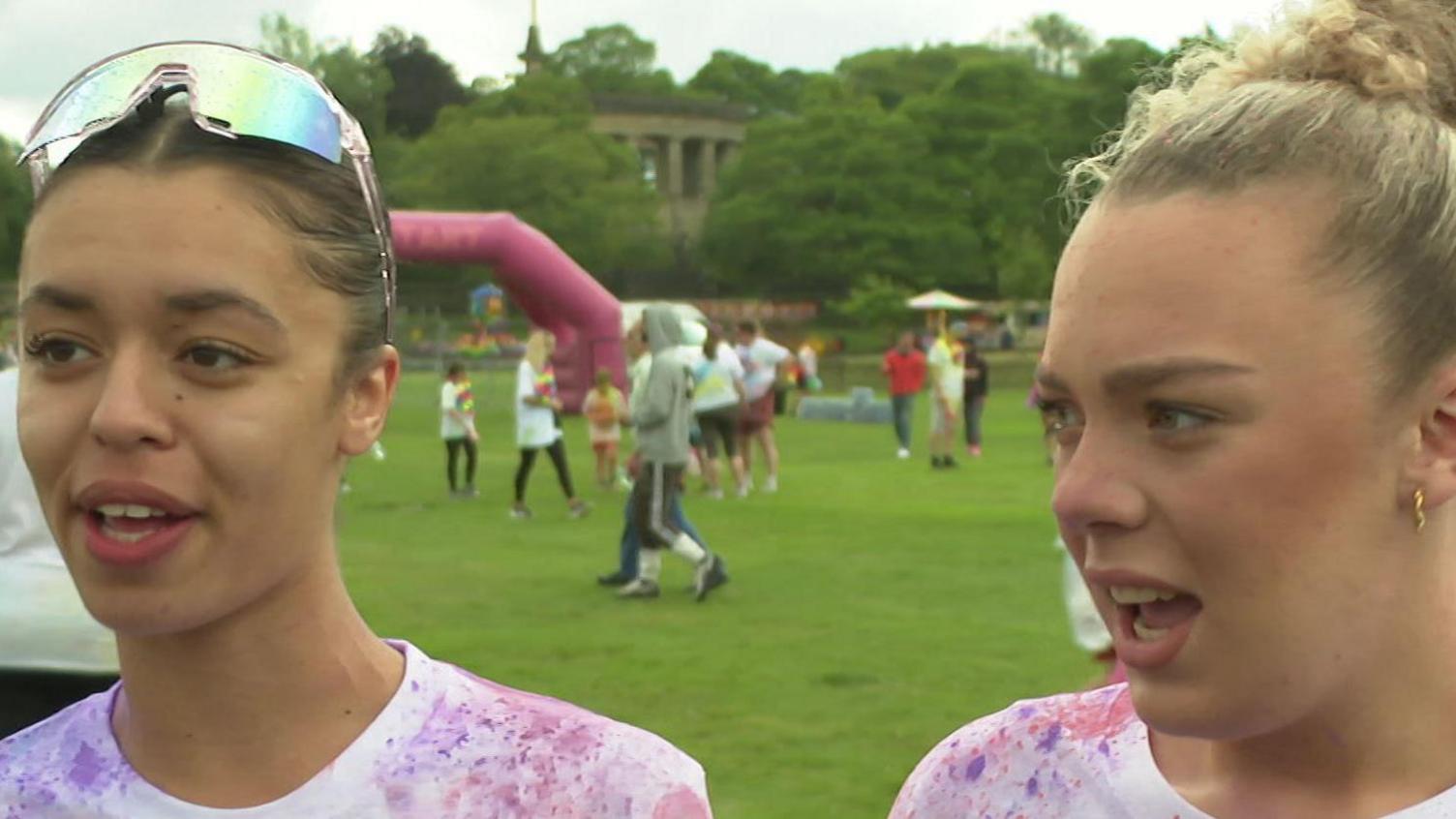 A young black woman with brown eyes and mirror sunglasses on her head in a white t-shirt stands next to a young blonde white girl with her hair pulled up into a knot.