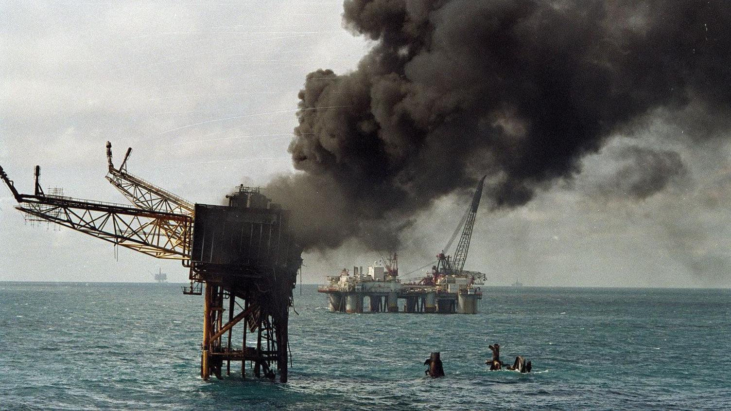 Thick smoke continues to billow from the wreck and a support platform can be seen in the background. The sea is calm.