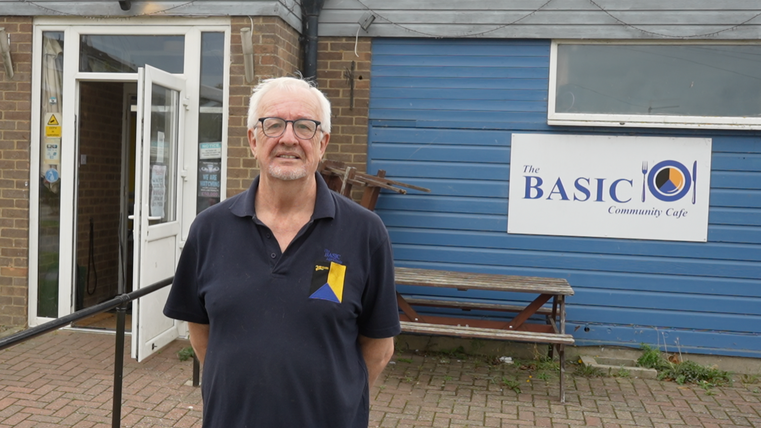 Graham Denny wearing a navy polo shirt and standing in front of the The Basic Community Cafe. He is looking into the camera.