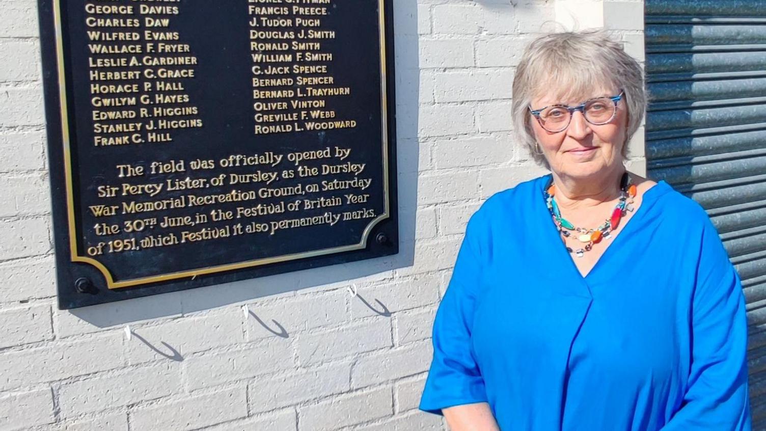 Julie Campbell wearing a blue shirt and stood next to a plaque with names of fallen soldiers on it