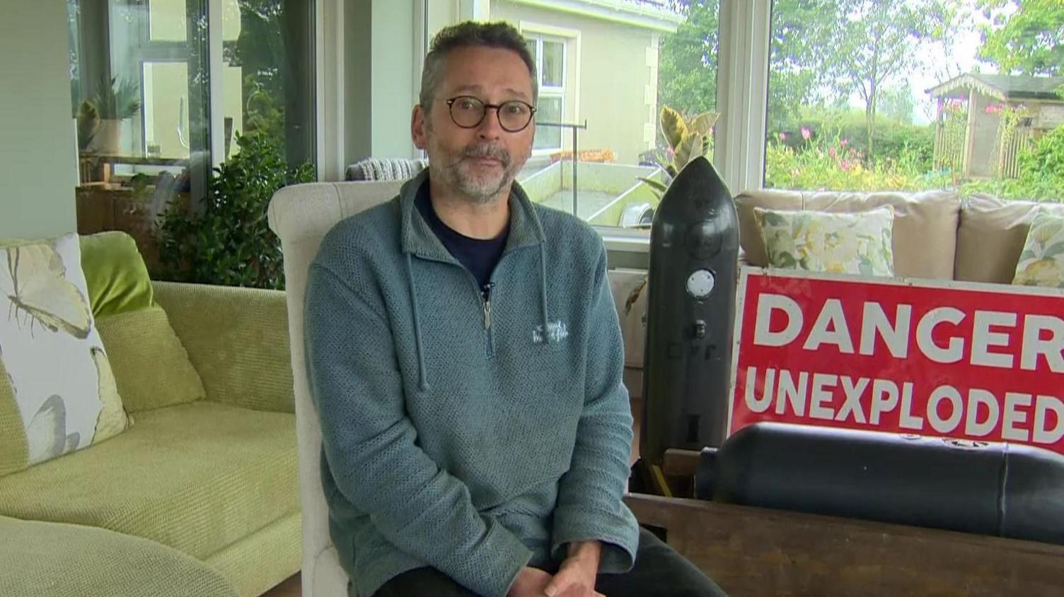 Jonny McNee sitting in his living room, in the background is two replica's similar to the bomb found in Newtownards. 