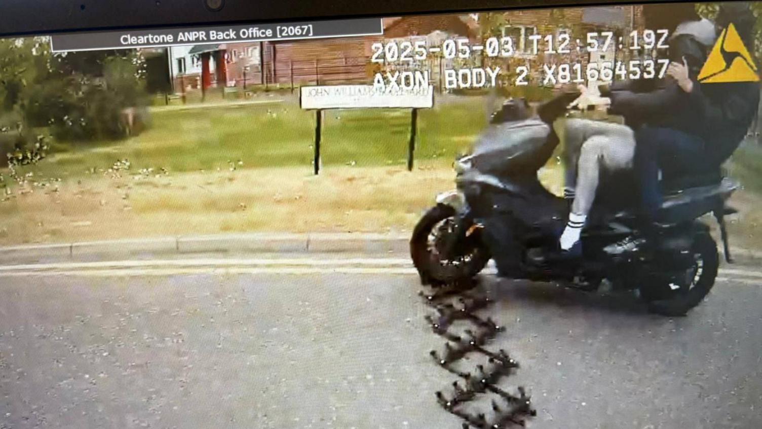 A still from a police body camera of the moped riders travelling over the stinger trap. The front wheel is directly above the spikes. There are two people on the moped, one in control and another holding on to him from behind.
