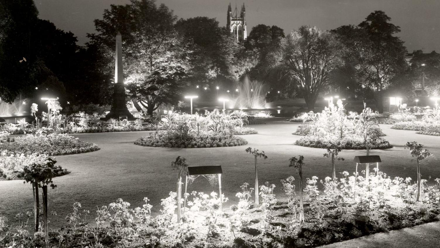 A black and white park scene with illuminated trees and bushes. In the background are fountains and the tower of Leamington Spa's All Saints Church.